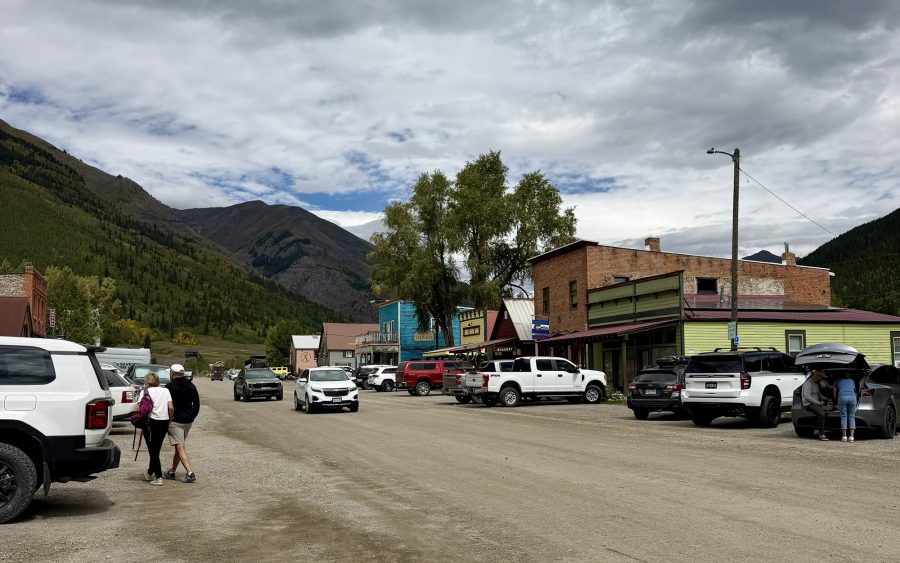 A street view of a small town with parked vehicles, shops, and mountains in the background, under a cloudy sky.