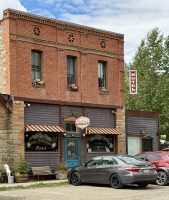 A historic brick building with a striped awning and a teal door, featuring large windows displaying decor. A sign reads 'Open' on the side. Several cars are parked in front.